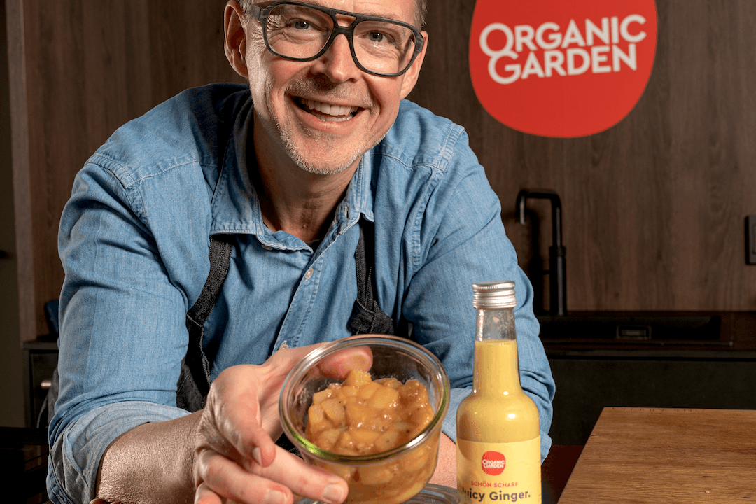 Holger Stromberg vor dem Logo von Organic Garden. Er hält ein Glas mit Apfel-Ingwer-Kompott in der Hand. Vor ihm steht eine Flasche Juicy Ginger Ingwersaft mit Apfelsüße